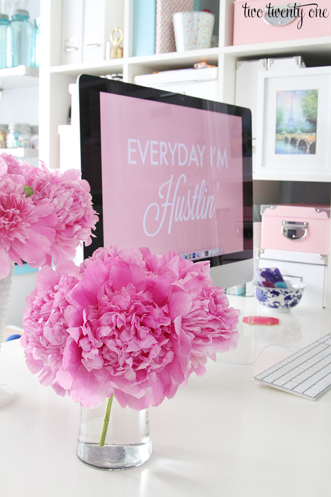 Fresh cut peonies in a clear vase setting in a white desk with a computer monitor in the background.