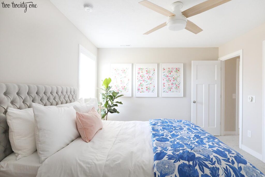 Bedroom with walls painted with Worldly Gray by Sherwin Williams. Bed with a gray upholstered headboard and blue floral comforter.