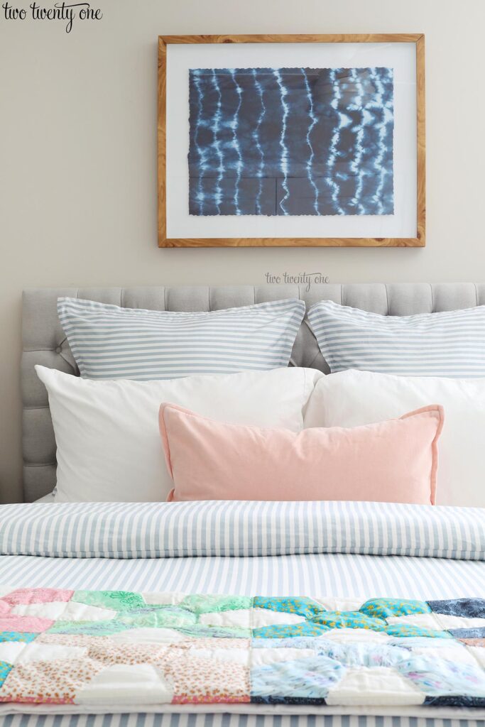 bed with vintage quilt, blue and white striped duvet, blue striped euro shams, white pillows, and light pink pillow. Shibori wall art above the bed. Wall color is Worldly Gray.