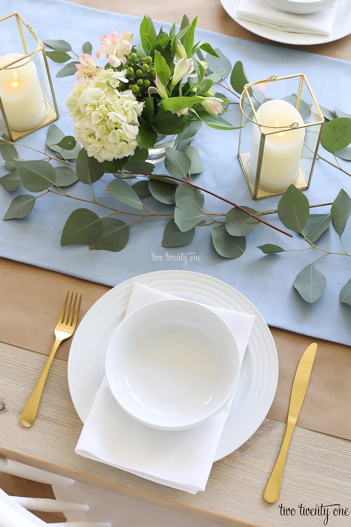 table set with blue table runner off-white candles in gold trimmer lanterns and flower arrangements of hydrangeas and greenery, white plate topped with a white napkin and white bowl with gold fork and knife