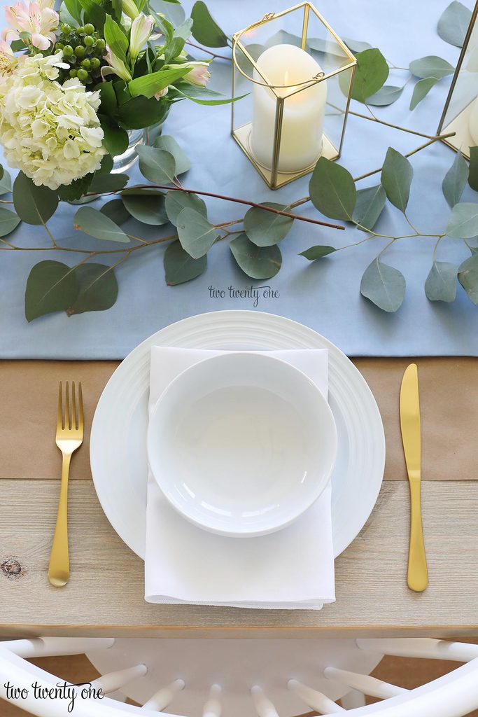 table set with blue table runner off-white candles in gold trimmer lanterns and flower arrangements of hydrangeas and greenery, white plate topped with a white napkin and white bowl with gold fork and knife