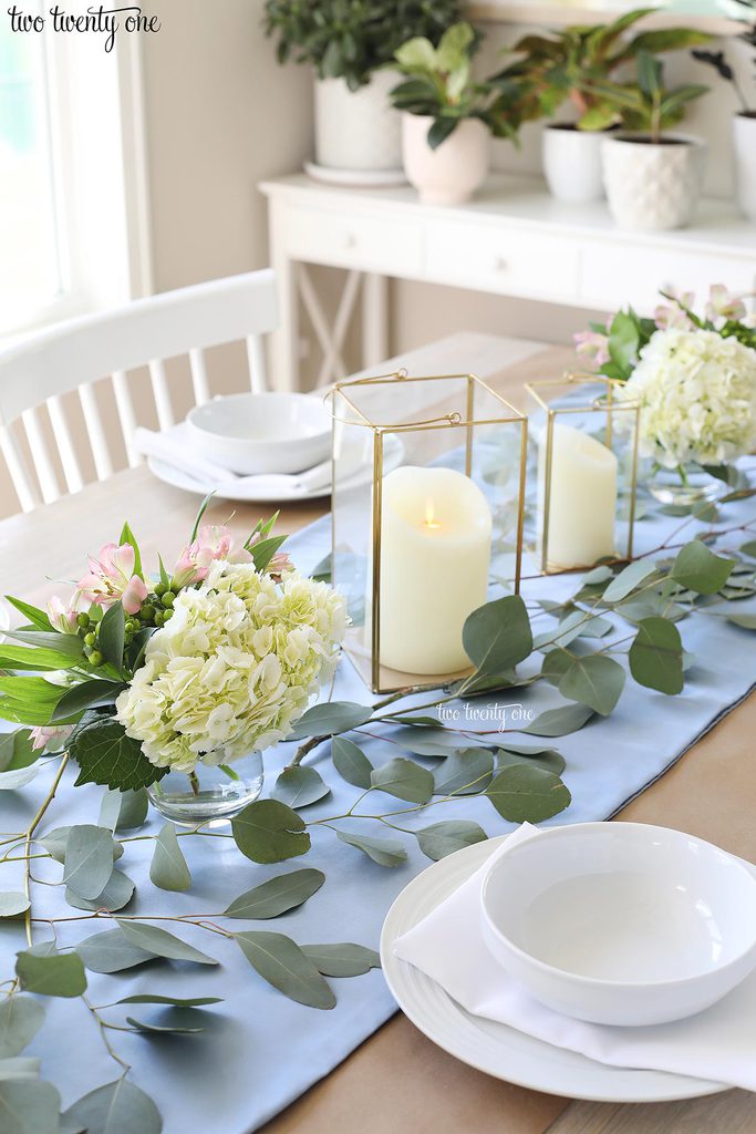 table set with blue table runner off-white candles in gold trimmer lanterns and flower arrangements of hydrangeas and greenery