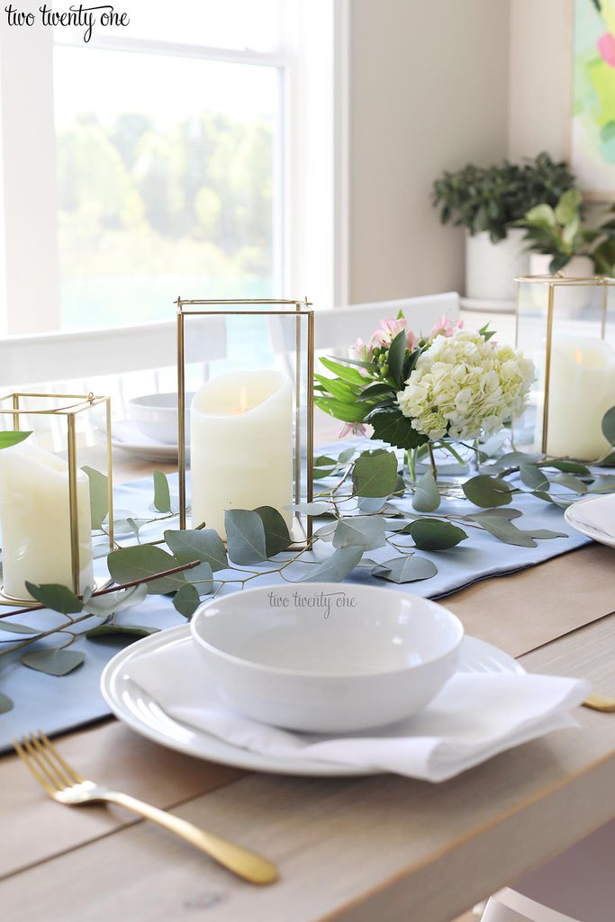 table set with blue table runner off-white candles in gold trimmer lanterns and flower arrangements of hydrangeas and greenery and white table setting with gold utensils