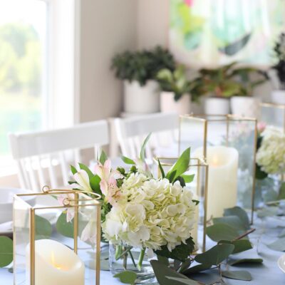 table set with blue table runner off-white candles in gold trimmer lanterns and flower arrangements of hydrangeas and greenery