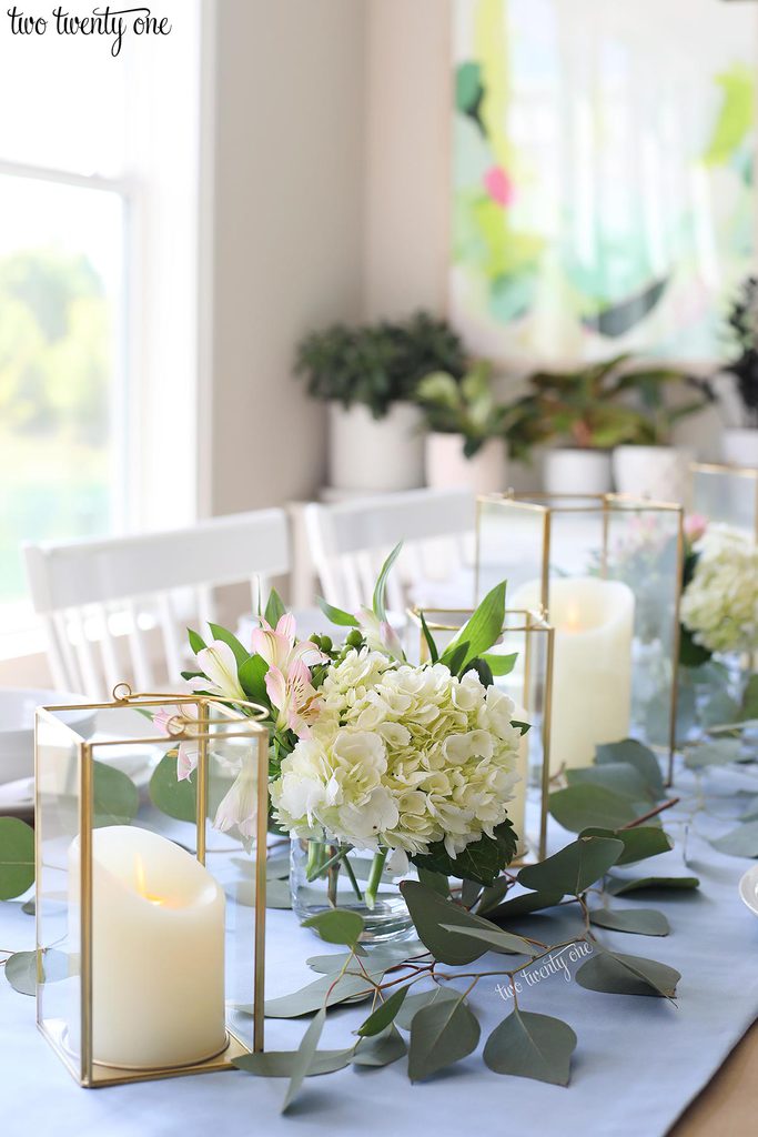 table set with blue table runner off-white candles in gold trimmer lanterns and flower arrangements of hydrangeas and greenery