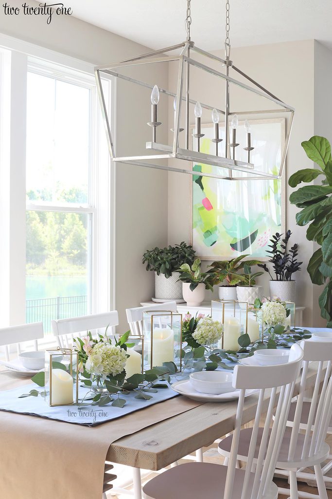 table set with blue table runner off-white candles in gold trimmer lanterns and flower arrangements of hydrangeas and greenery