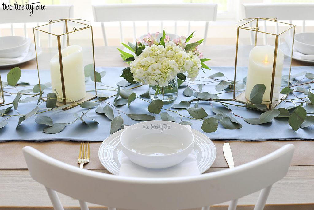 table set with blue table runner off-white candles in gold trimmer lanterns and flower arrangements of hydrangeas and greenery and white table setting with gold utensils