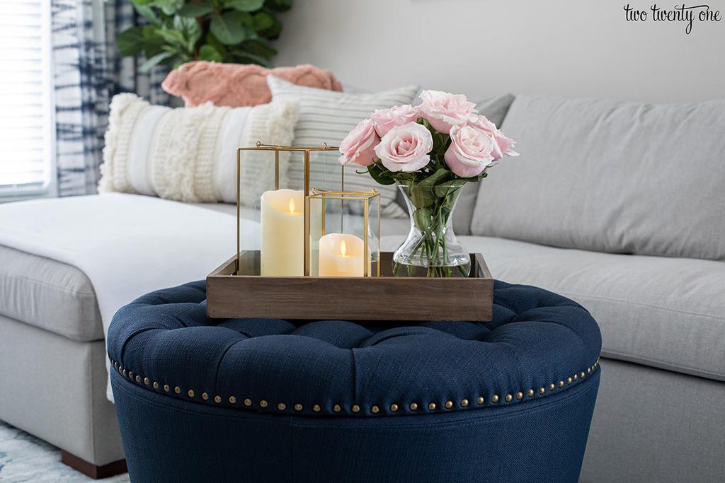 navy ottoman with tray on top. Tray has two lanterns with candles in them and a vase filled with pink roses