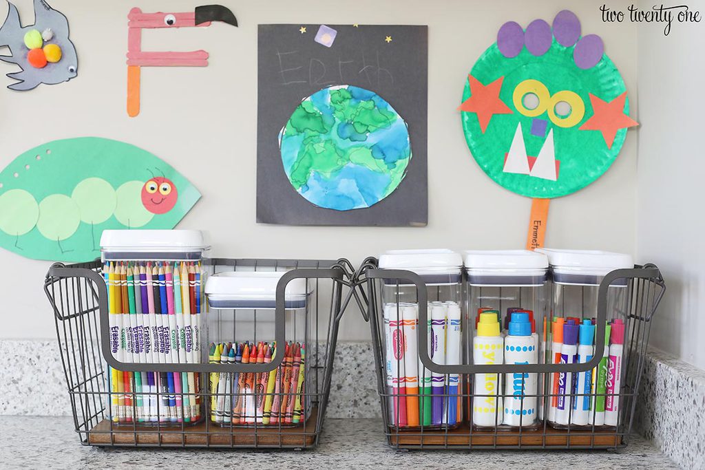 Markers, crayons, colored pencils in clear storage containers. The containers are inside wire and wood baskets.