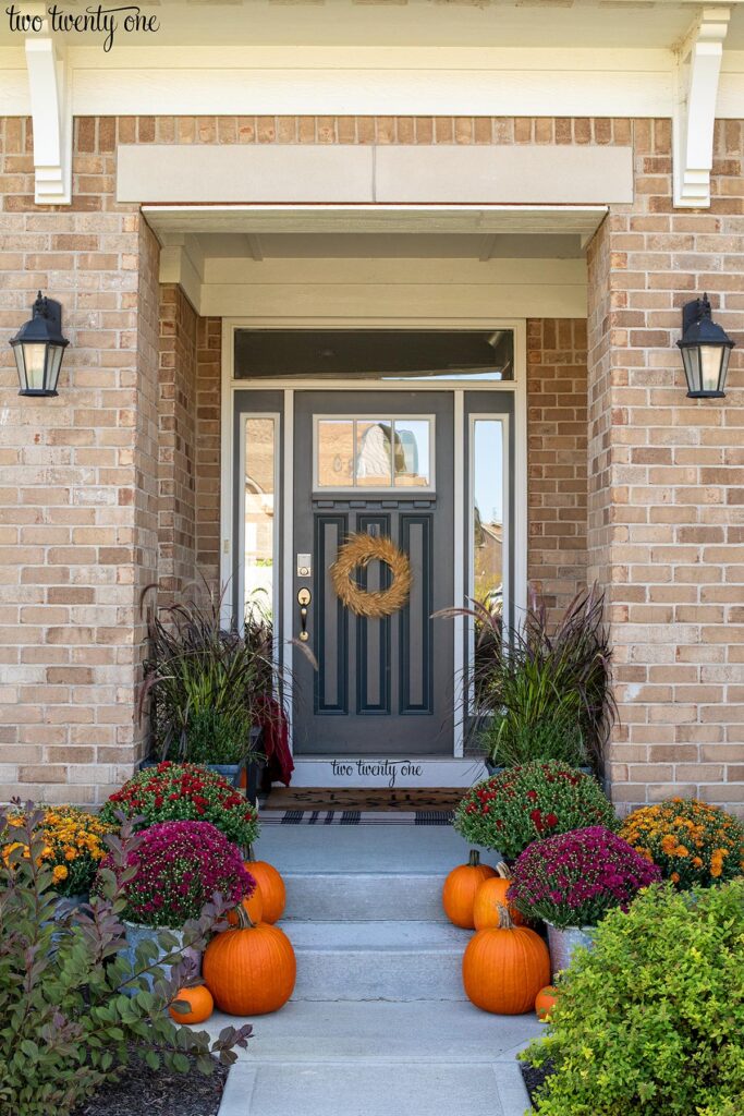 Front porch decorated for fall with mums, pumpkins, ornamental grass, and dried wreath on the front door