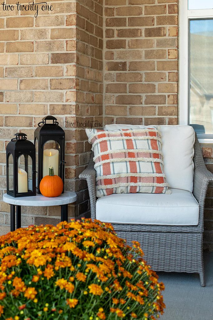 front porch chair with cream and orange plaid throw pillow. Side table with two black lanterns with candles and small pumpkin