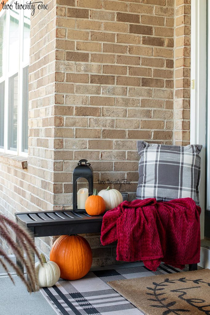Black bench with dark red throw blanket, gray and white plaid pillow, black lantern with candle, and pumpkins