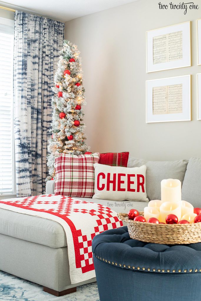 flocked christmas tree decorated with red ornaments behind a gray chaise sofa with holiday pillows and a navy ottoman with a basket filled with candles and red ornaments