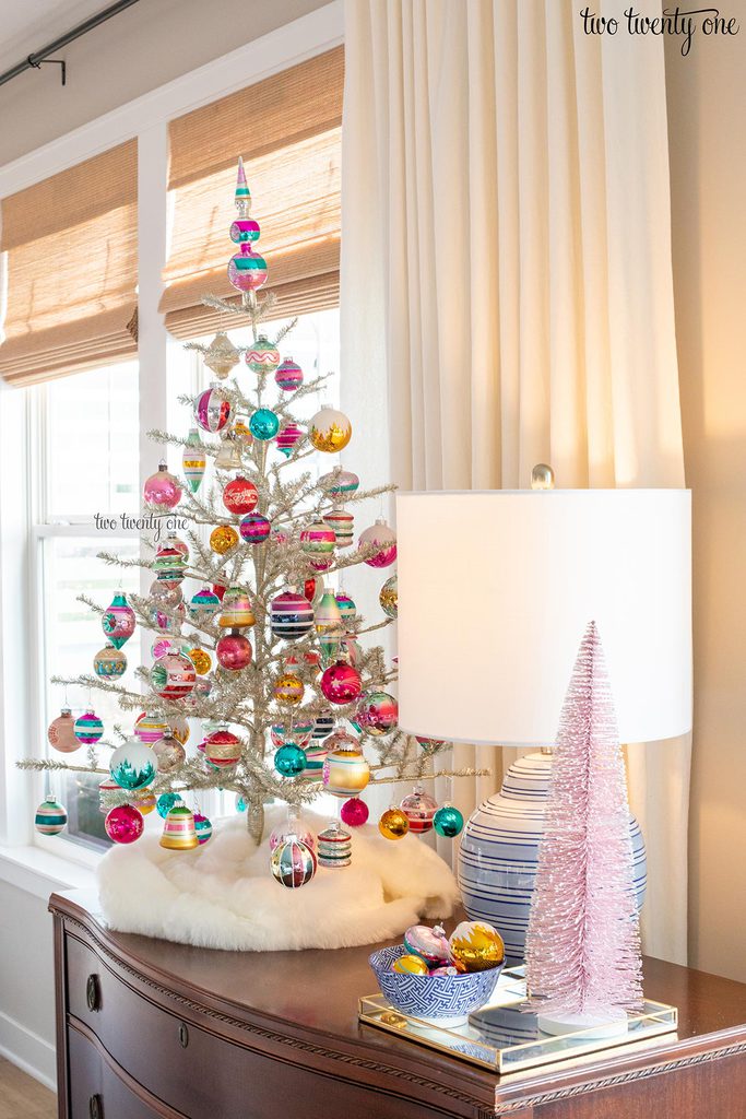 colorful glass ornaments on a silver tinsel christmas tree with a lamp, pink bottle brush tree, and bowl of ornaments