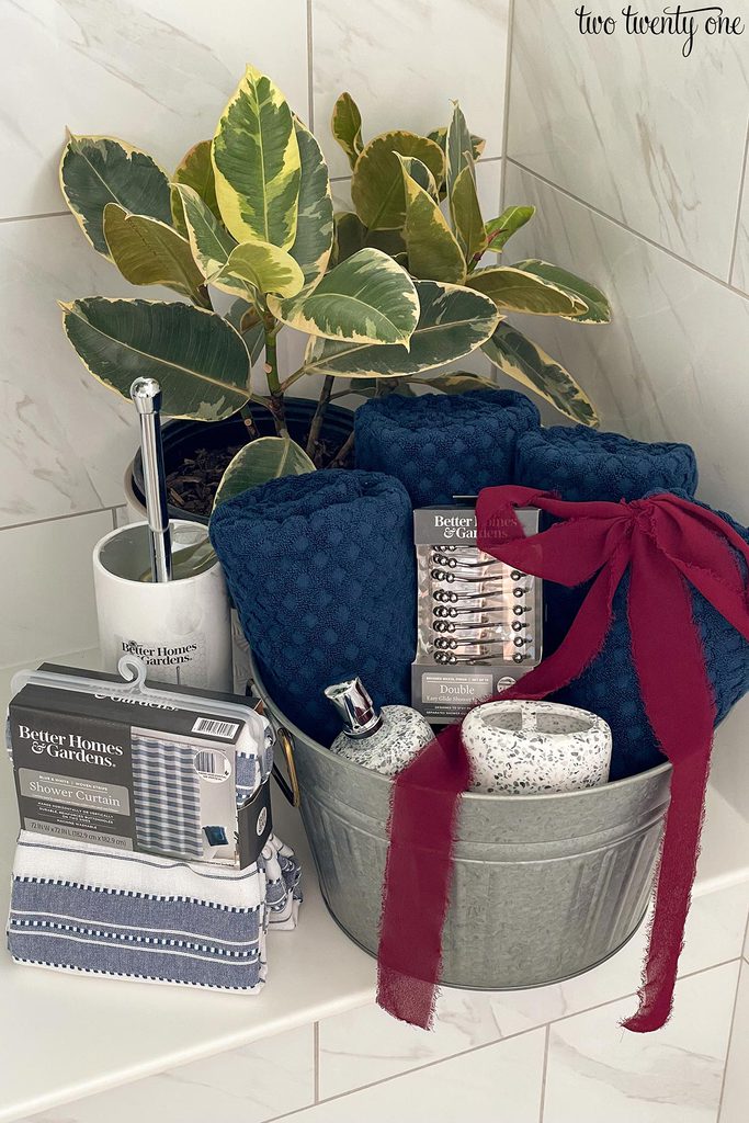 A galvanized bucket with four navy towels, lotion dispenser, toothbrush holder, and shower curtain hooks. A shower curtain and toilet brush are to the side of the bucket.