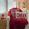 A white chair with a red blanket draped on it with two pillows on top-- one plaid and one reads CHEER. A small table is to the side of the chair, topped with red ornaments and a glass hurricane with a red candle inside. A floorlamp and undecorated christmas tree are in the background.