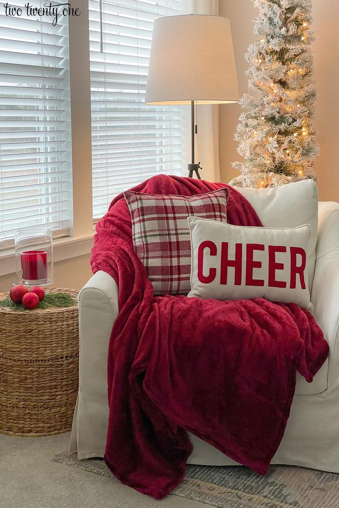 A white chair with a red blanket draped on it with two pillows on top-- one plaid and one reads CHEER. A small table is to the side of the chair, topped with red ornaments and a glass hurricane with a red candle inside. A floorlamp and undecorated christmas tree are in the background.