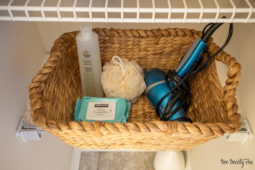Body wash, facial wipes, loofah, and hair dryer inside a woven basket on a wire shelf in a linen closet.