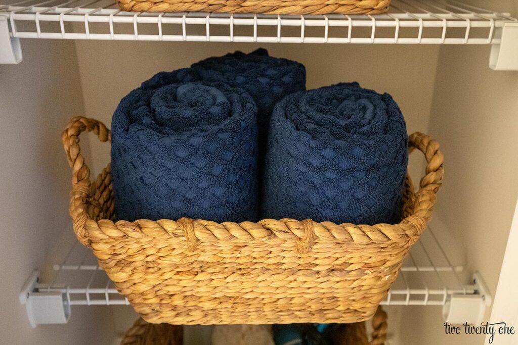 Navy bath towels rolled up inside a woven basket on a wire shelf in a linen closet.