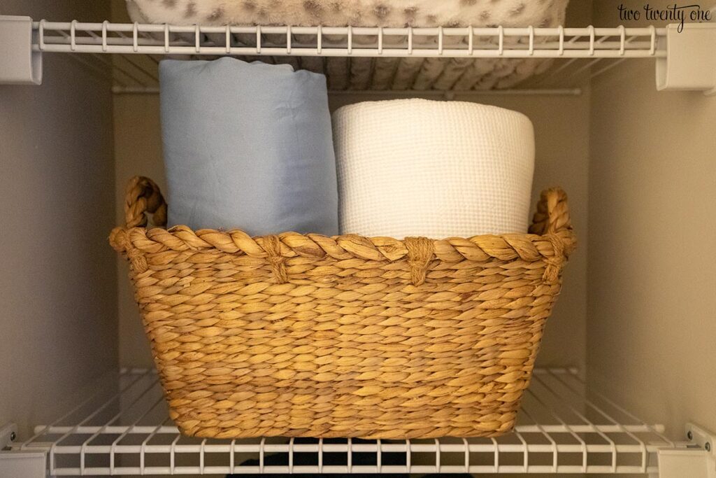 Sheets and a blanket inside a woven basket on a wire shelf in a linen closet.