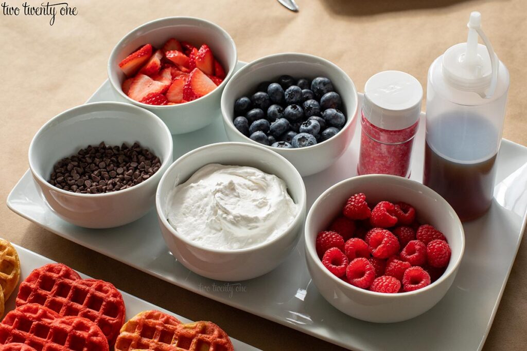 strawberries, blueberries, sprinkles, syrup, mini chocolate chips, whipped cream, and raspberries in separate white bowls