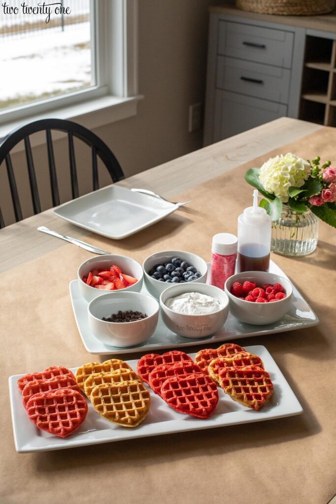 platter of heart shaped waffles and a platter with waffle toppings on a table for a valentine's day brunch 