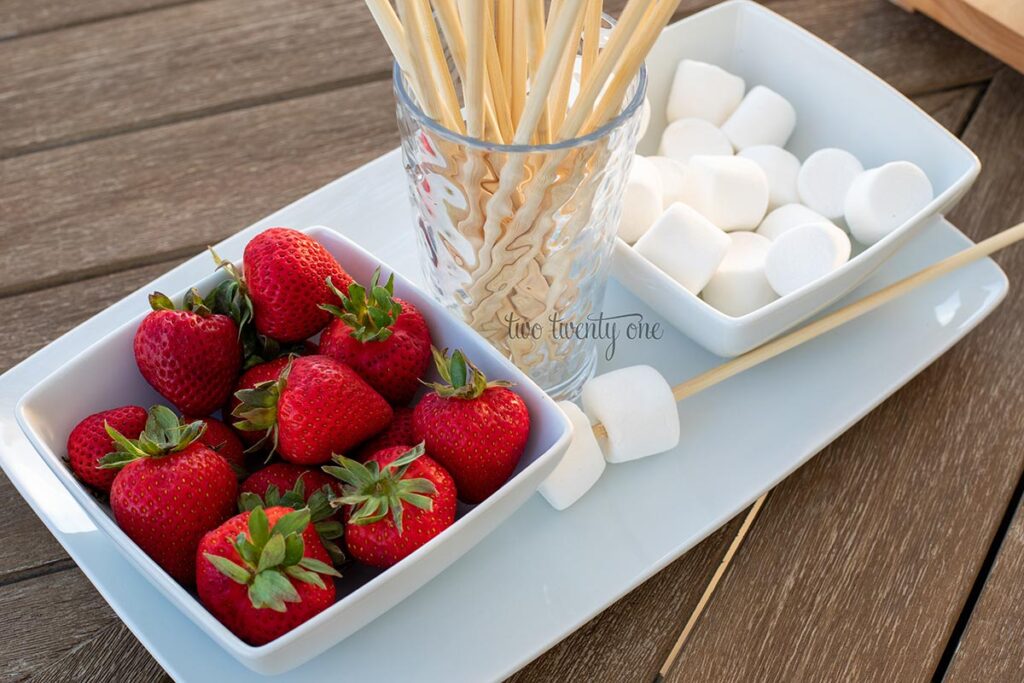 smores station strawberries in a bowl, wooden skewers in a cup, and marshmallows in a bowl