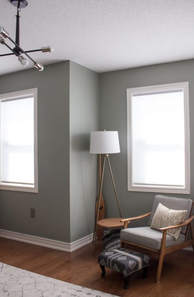 Bedroom with white painted ceiling, trim and baseboards. The wall color is Benjamin Moore Oil Cloth, a muted green. A lamp sits next to an end table and modern-looking chair.