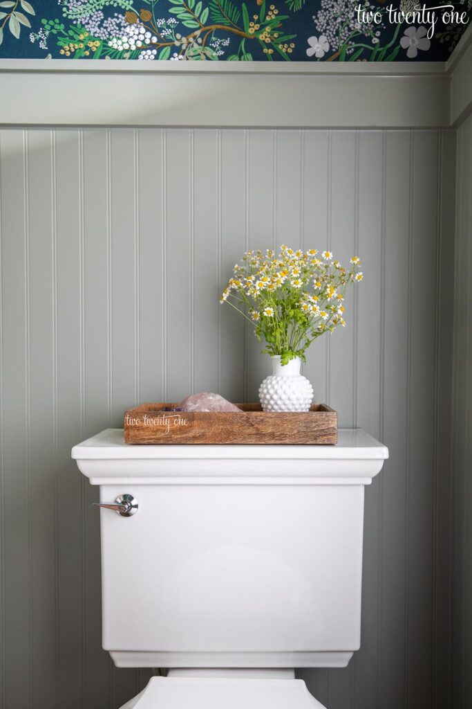 Benjamin Moore Oil Cloth paint on beadboard paneling in a bathroom with botanical wallpaper above the paneling. The top portion of a toilet is shown with a tray containing flowers and rose quartz on top of the tank.