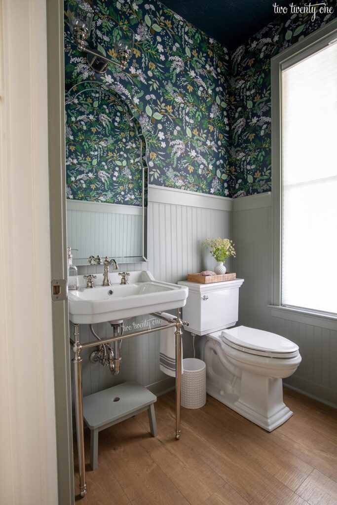 A powder room with botanical wallpaper on the top portion of the walls and beadboard paneling painted with Benjamin Moore Oil Cloth on the bottom portion. There is a white sink, white toilet, white trash can, mirror, step stool, and window.