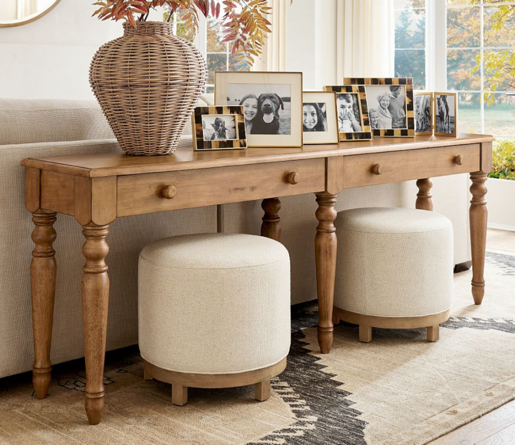 Pottery Barn Heritage wood stained sofa table sits behind a tan sofa. Picture frames with black and white photos set on top of the sofa table with a large woven vase. Two cylinder-shaped, upholstered stools are stored underneath the console table.